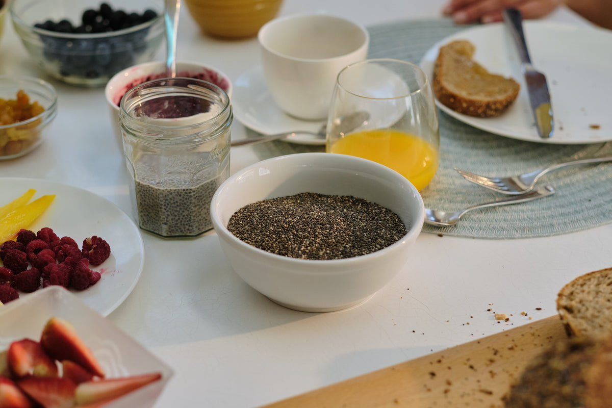 A breakfast table with a bowl of chia seeds, a jar of soaked chia, orange juice, berries and bread