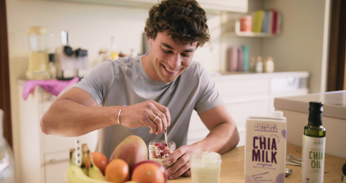 A man stirs a jar of chia pudding at a kitchen counter, beside a carton of Seeds of Wellness chia milk and a bottle of chia oil