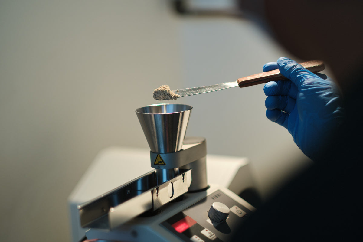 A gloved hand holds a spatula of chia fiber powder over a steel funnel in a lab