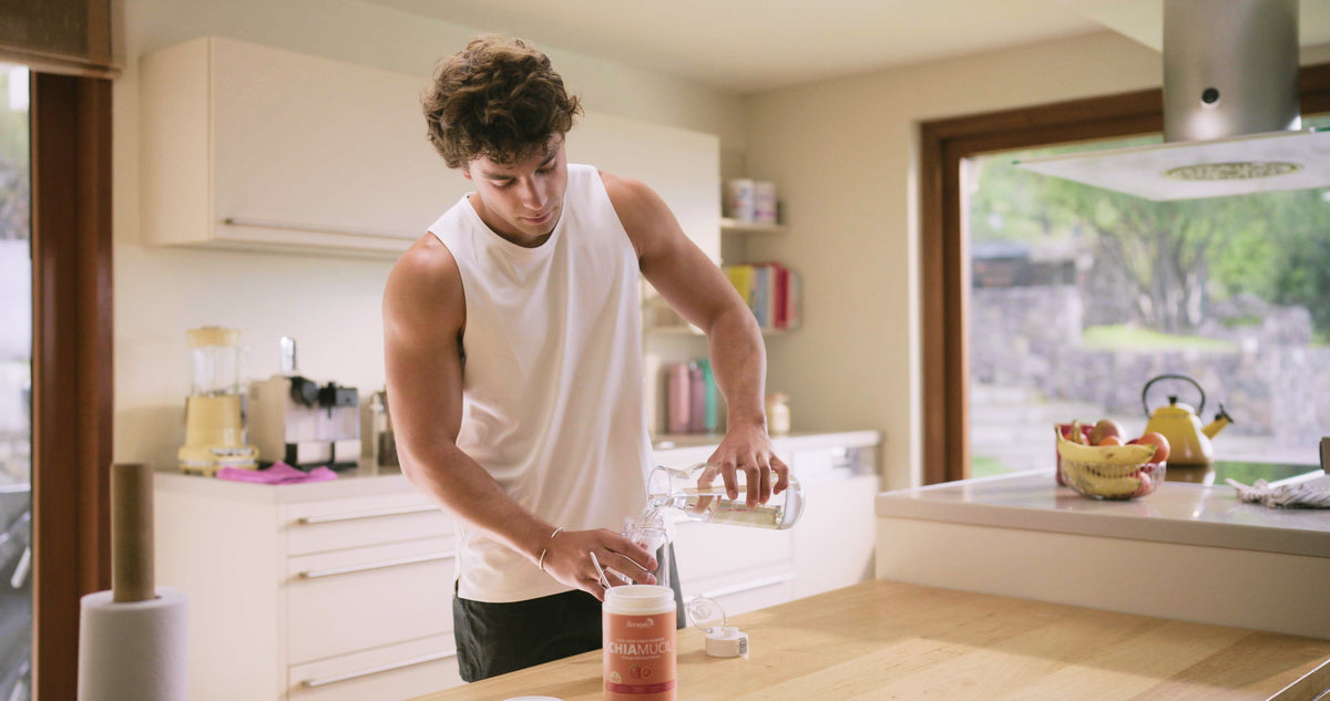 A man pours water into a glass in a bright kitchen, with a tin of ChiaMucil Orange Flavored on the counter